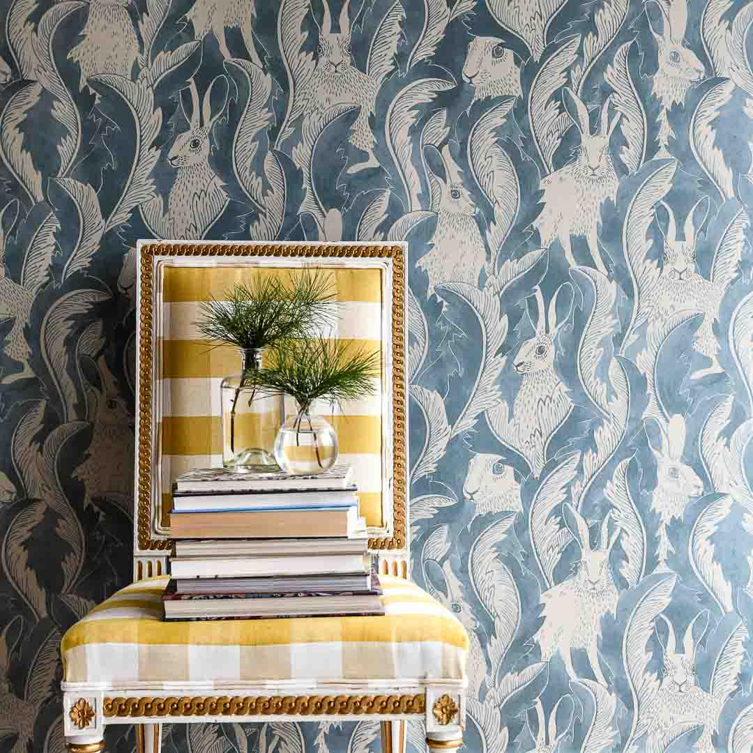 Chair stacked with books and glass vases in front of smokey blue wallpaper