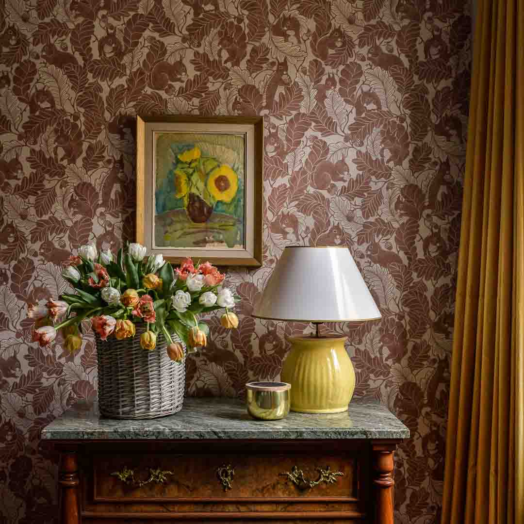 Decorative room interior with floral arrangement, lamp, and painting on patterned wall.
