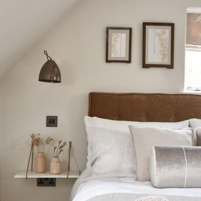 A styled bedroom corner featuring a bed with white and light gray bedding, a brown upholstered headboard, a small floating shelf with two vases, a wall-mounted dome lamp, and two framed botanical prints on the wall.