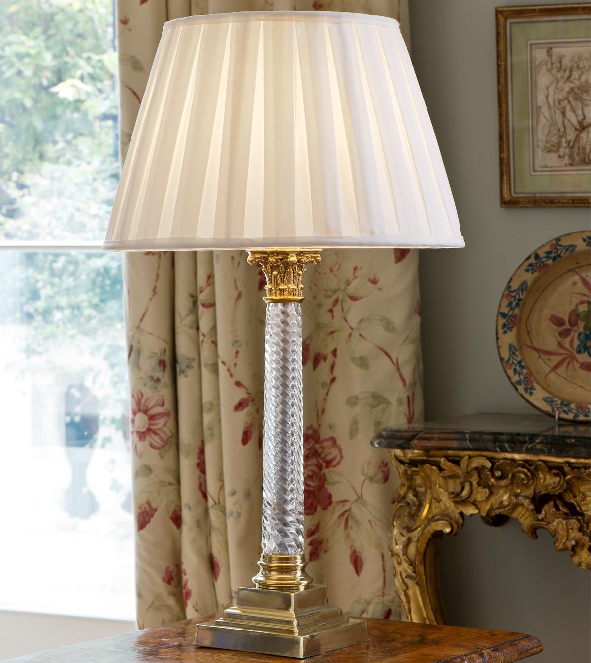 Glass and brass column light with pleated silk cream  lampshade in front of floral curtain